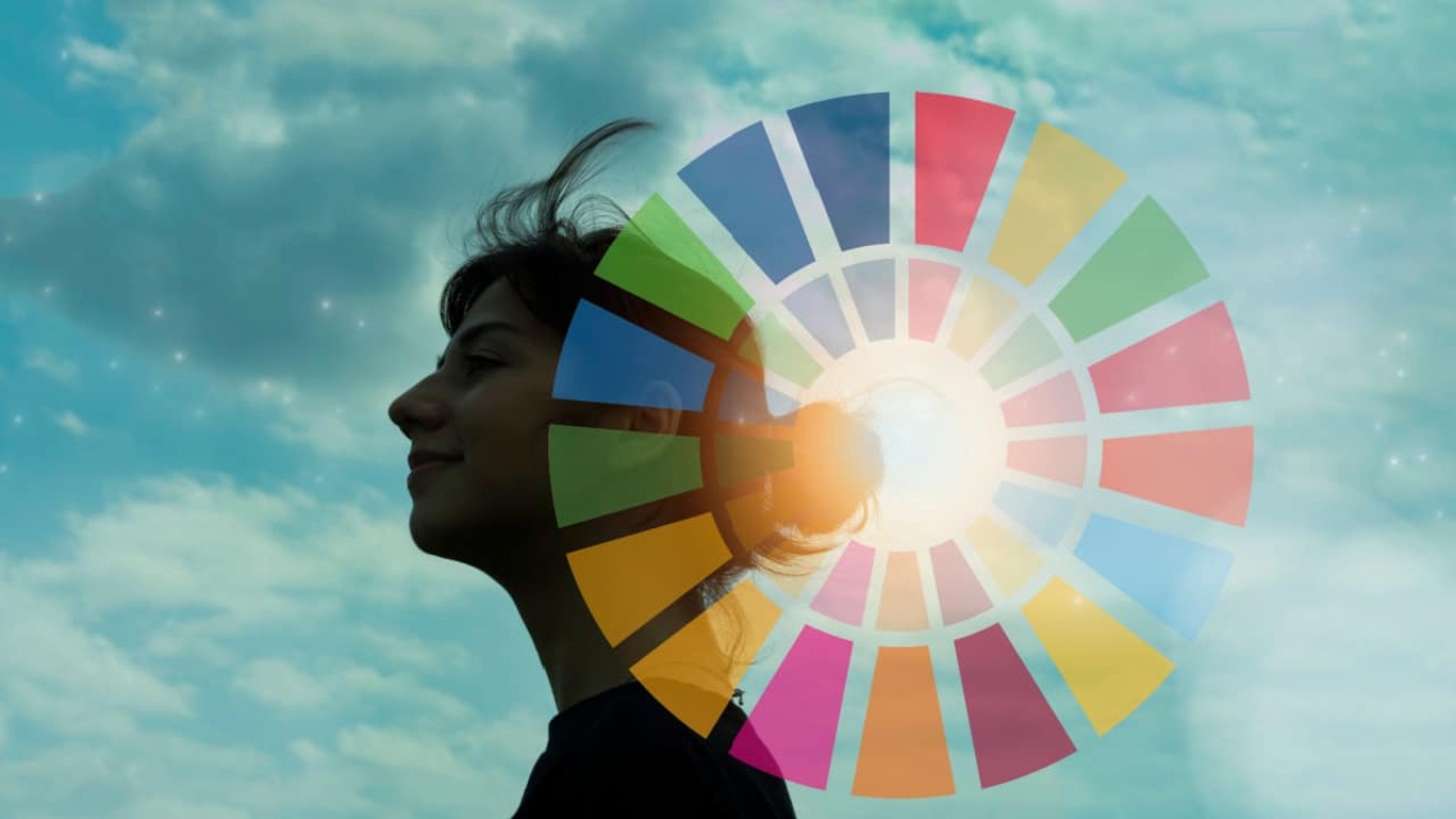 Mulher branca jovem com céu azul e nuvens ao fundo e um círculo representando os Objetivos de Desenvolvimento Sustentável sobreposto à imagem.