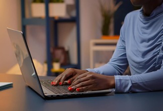 Mulher negra sentada a mesa, digitando num laptop, com unhas pintadas de vermelho.