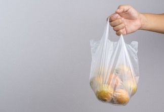 Sobre fundo cinza, uma mão masculina segura uma sacola plástica transparente com laranjas.