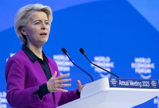 Mulher branca, com cabelos curtos e loiros claros, usando terno púrpura com blusa preta embaixo. Ela fala em palanque, com fundo azul atrás do Fórum Econômico Mundial.