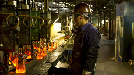 Fábrica de vidros da Owens-Illinois. Na imagem, um homem usa equipamento de segurança e observa a produção de algumas garrafas de vidro.