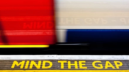 Sinal com os dizeres "Mind the Cap" escrito em amarelo com trem em movimento ao fundo